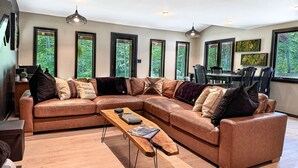 Un salon moderne avec un canapé d’angle, une table basse en bois et de grandes fenêtres donnant sur un paysage verdoyant.