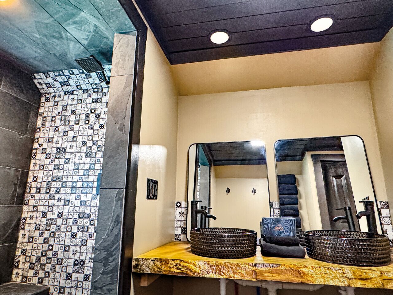 Un baño con pared de azulejos, dos lavamanos con recipientes negros y un espejo sobre los lavamanos.