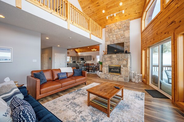 A spacious living area with a high ceiling, a stone fireplace, a wooden balcony, and a kitchen in the background. - Chic Rustic Log Cabin Near Lake w/ Fire Pit (Oakland)