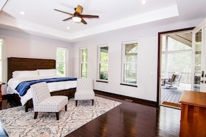 Une chambre dotée d’un grand lit, de deux fauteuils à motifs, d’un ventilateur de plafond et offrant une vue sur l’extérieur grâce à de grandes fenêtres.