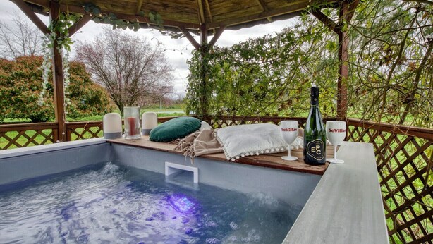 Een hottub met een houten terras, omringd door begroeiing en een pergola.