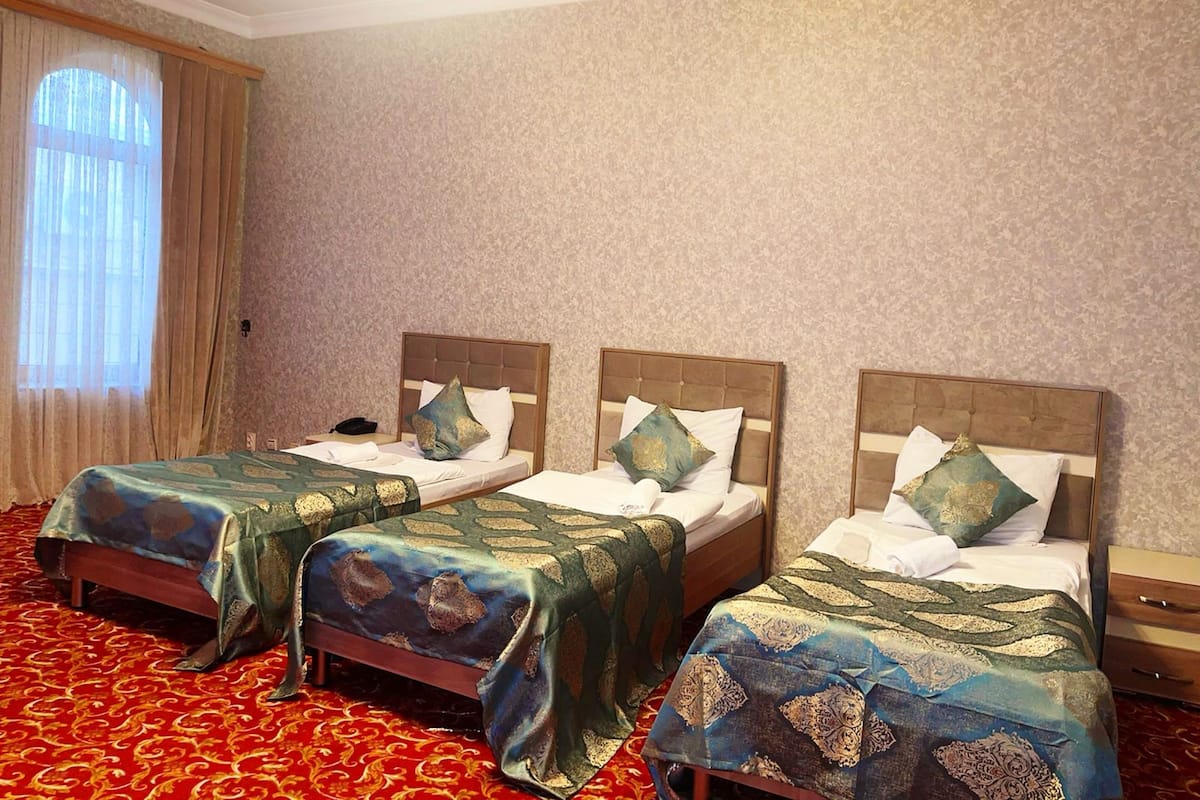 A hotel room with three single beds, each with a wooden headboard and a patterned bedspread. The room has a red carpet and a large window with curtains.