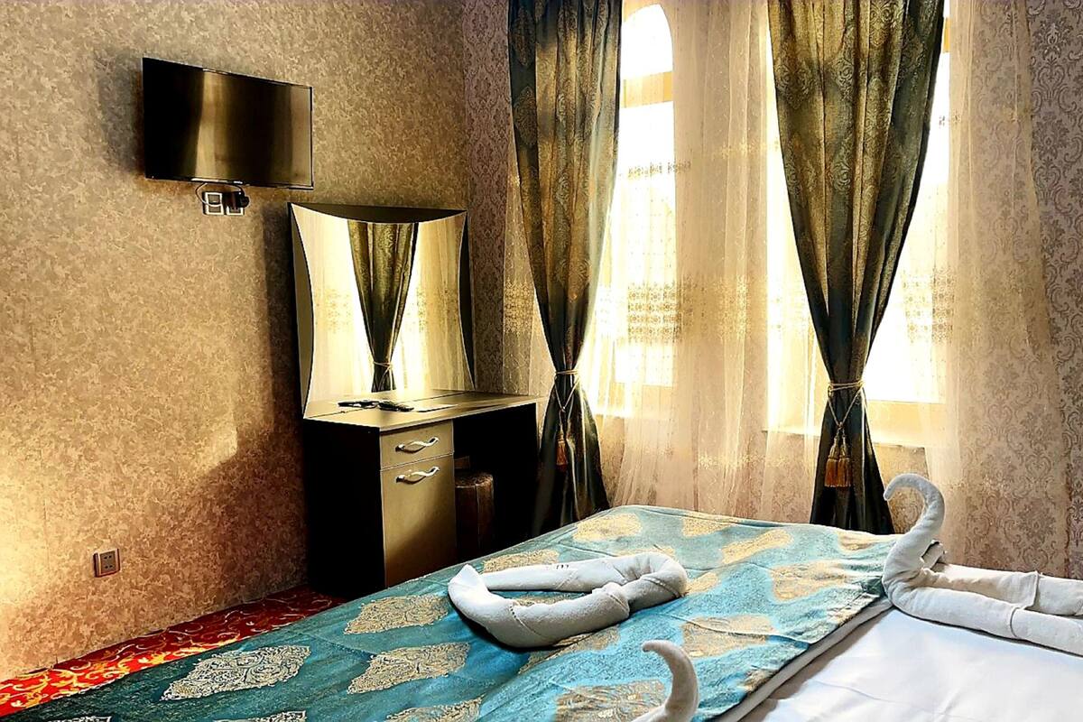 A hotel room with a bed, a TV mounted on the wall, a desk with drawers, and curtains on the windows.
