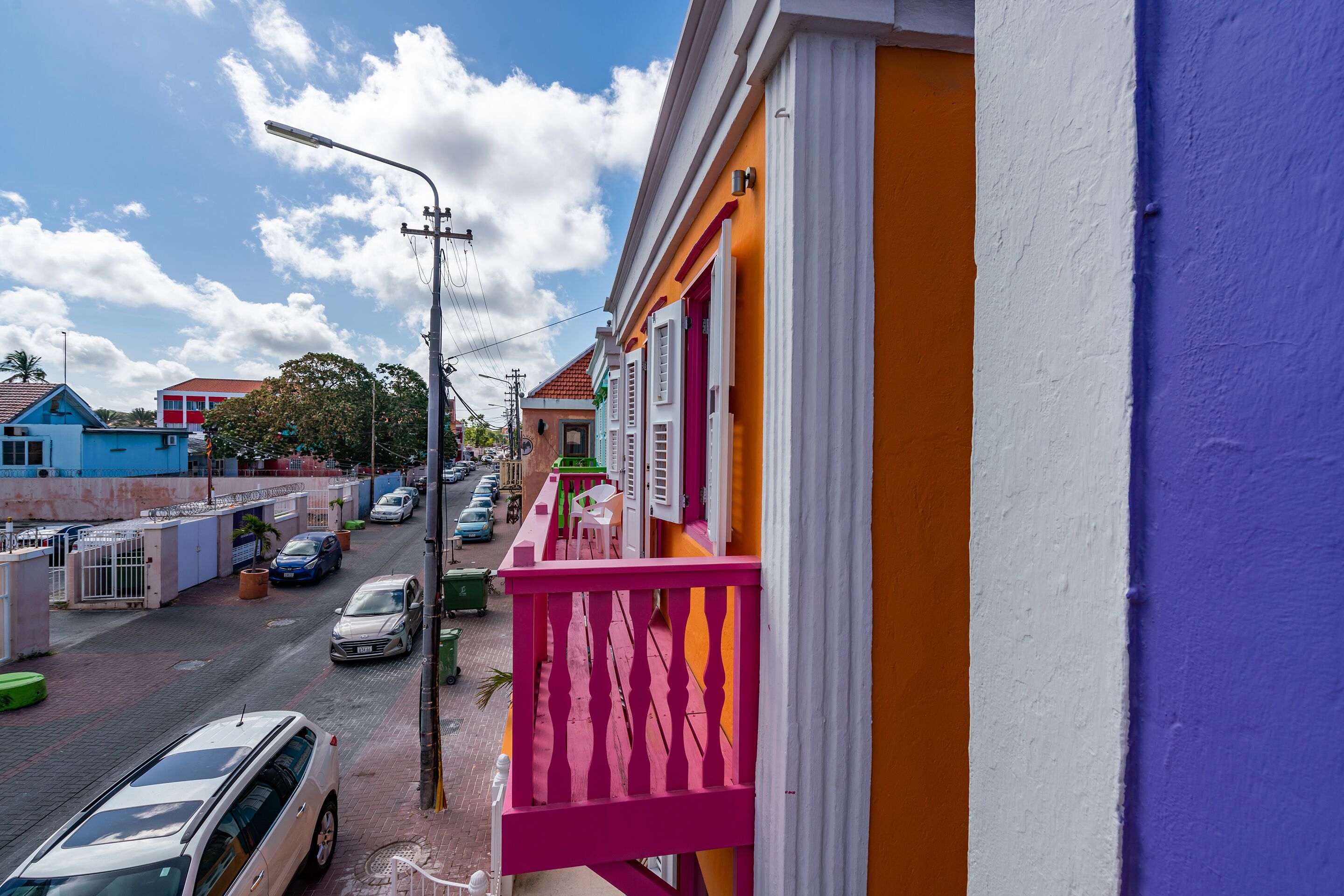 A street view with colorful buildings, a balcony with a pink railing, and parked cars.