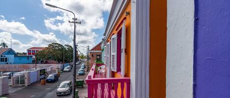 A street view with colorful buildings, a balcony with a pink railing, and parked cars.