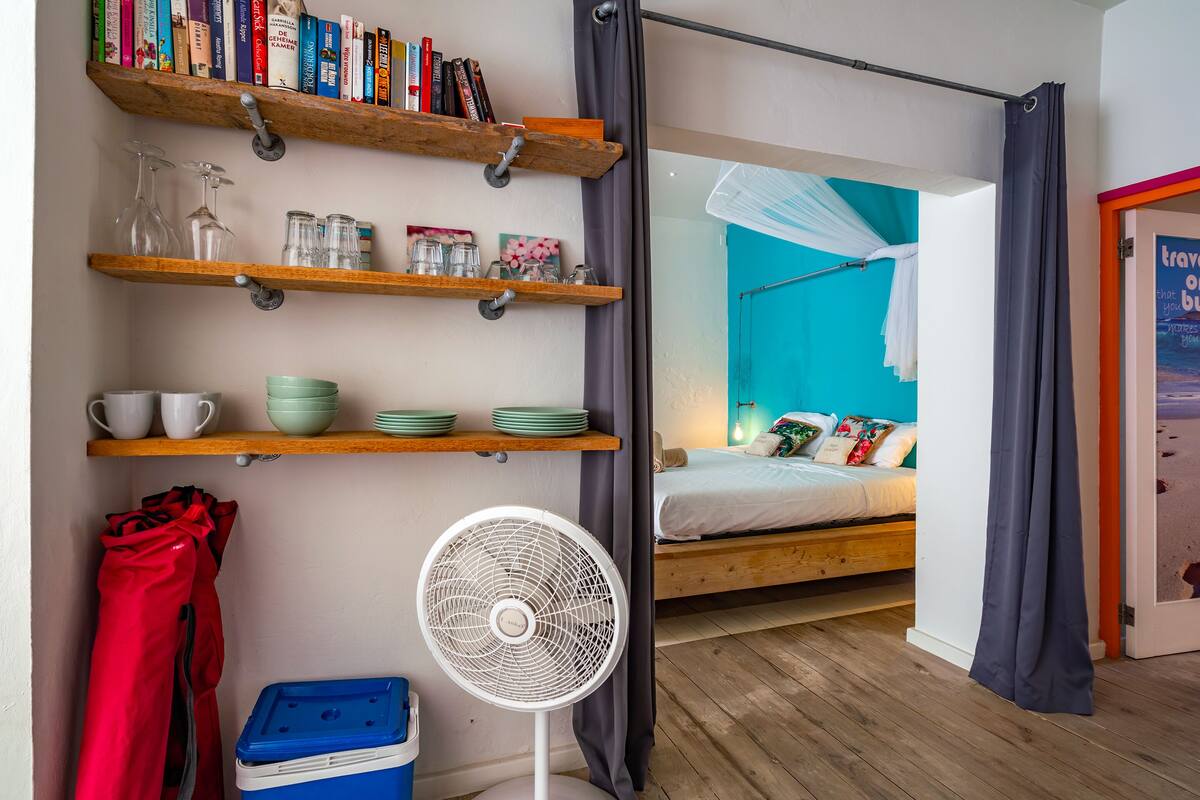 A room with a bed, a fan, shelves with books and kitchenware, and a view of another room with a bed and a blue wall.