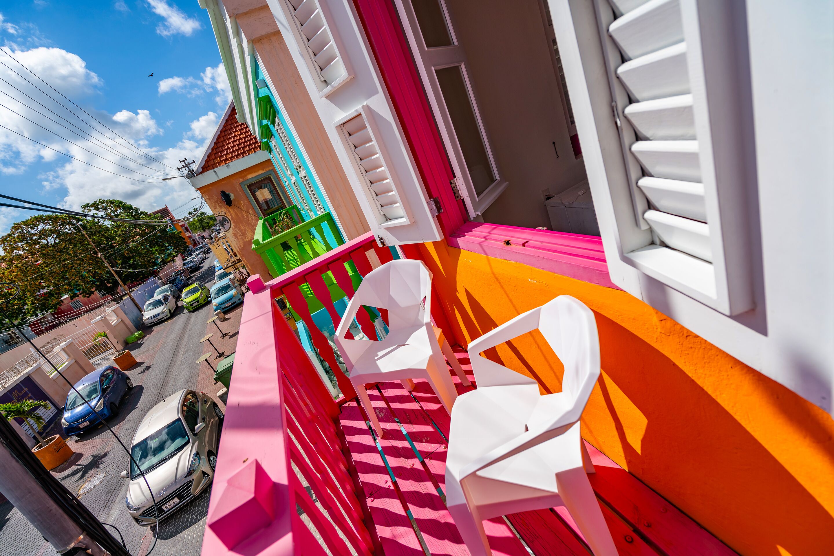 Un balcon avec des balustrades colorées, une chaise blanche et une vue sur les voitures garées et les immeubles.