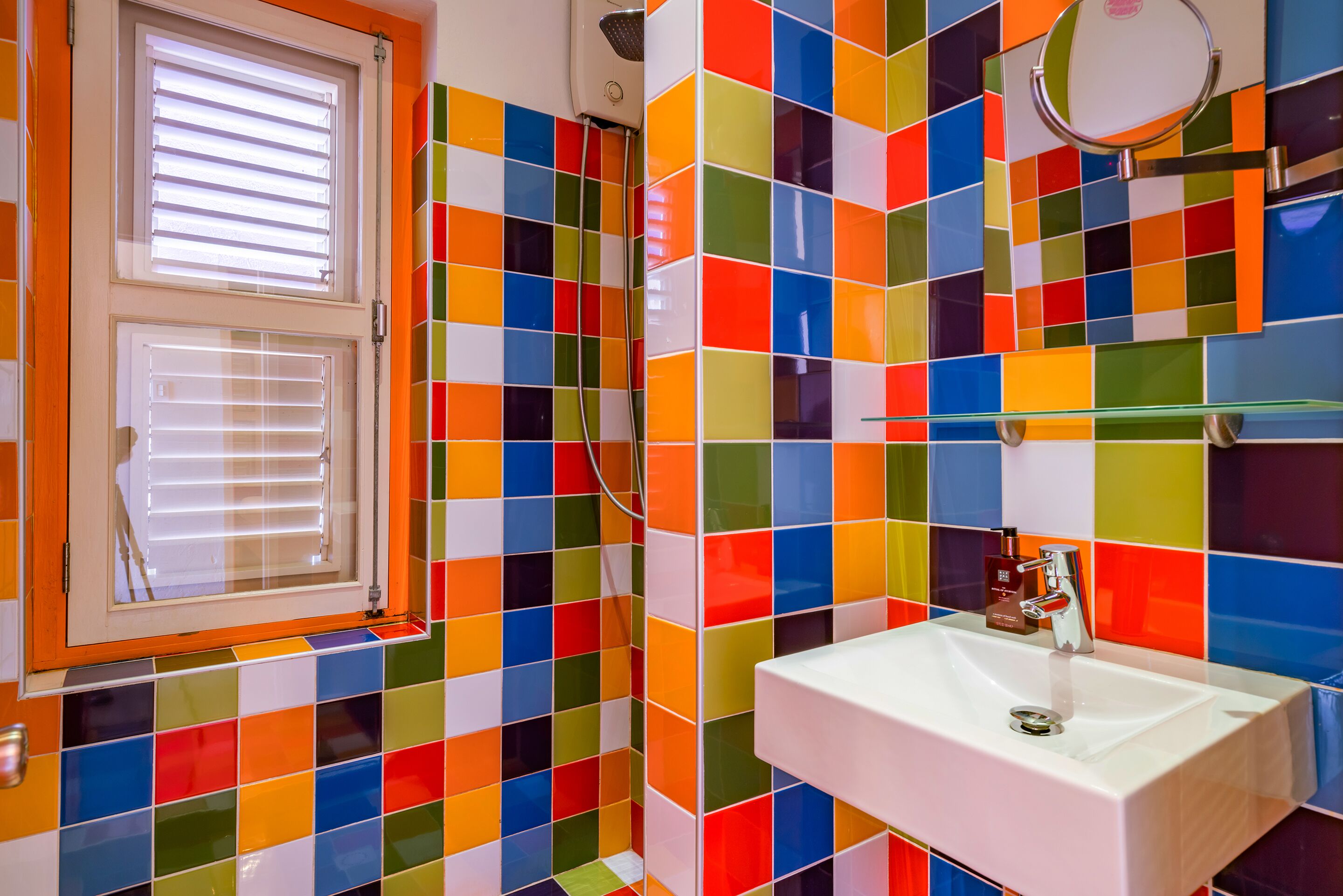 Casa de banho com azulejos coloridos, uma pia branca e uma janela com persianas.