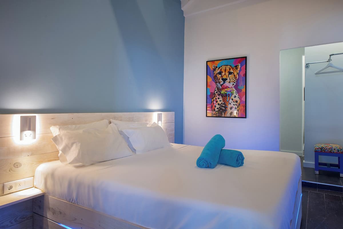 A neatly made bed with white linens and two rolled towels, a wall-mounted painting of a leopard, and a bedside table with a lamp.