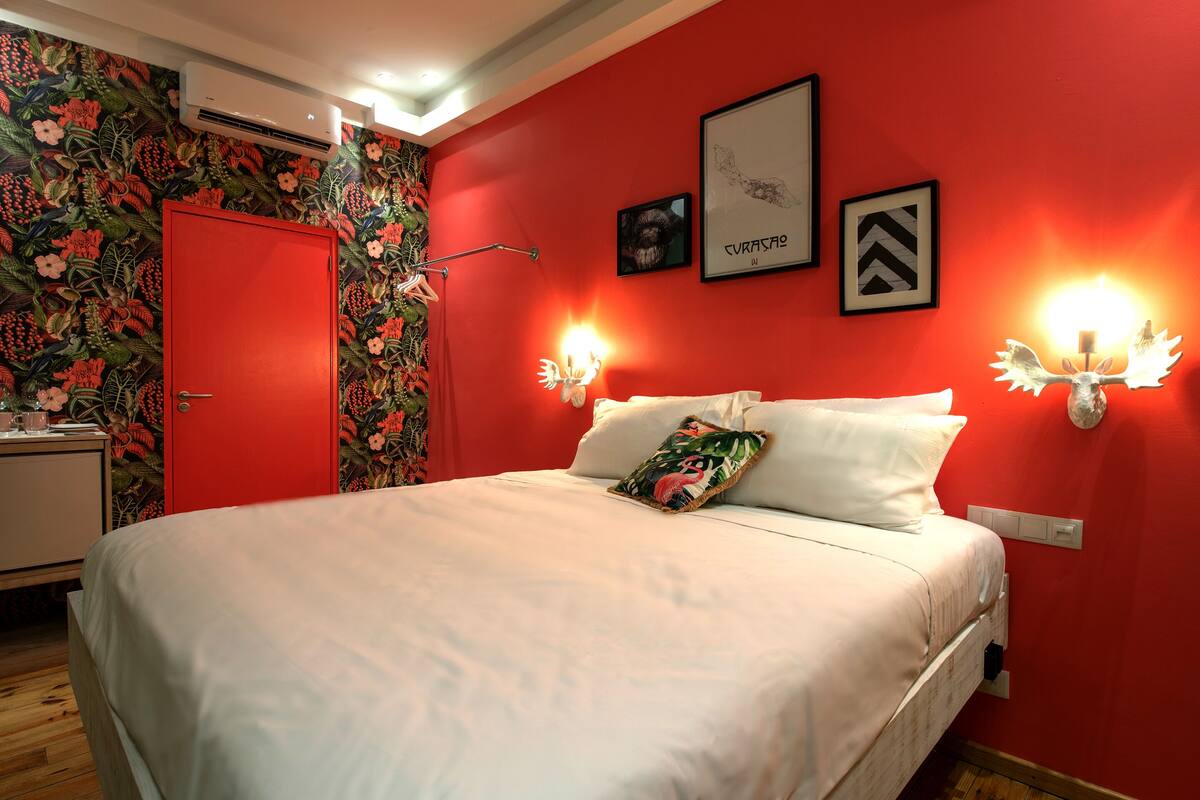A bedroom with a red wall, a floral wallpaper on one side, a white bed, and two wall-mounted lamps.