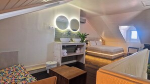 Un pequeño y acogedor dormitorio en el ático, con techo inclinado, tocador de madera empotrado con espejo, una cama individual y una pequeña mesa.