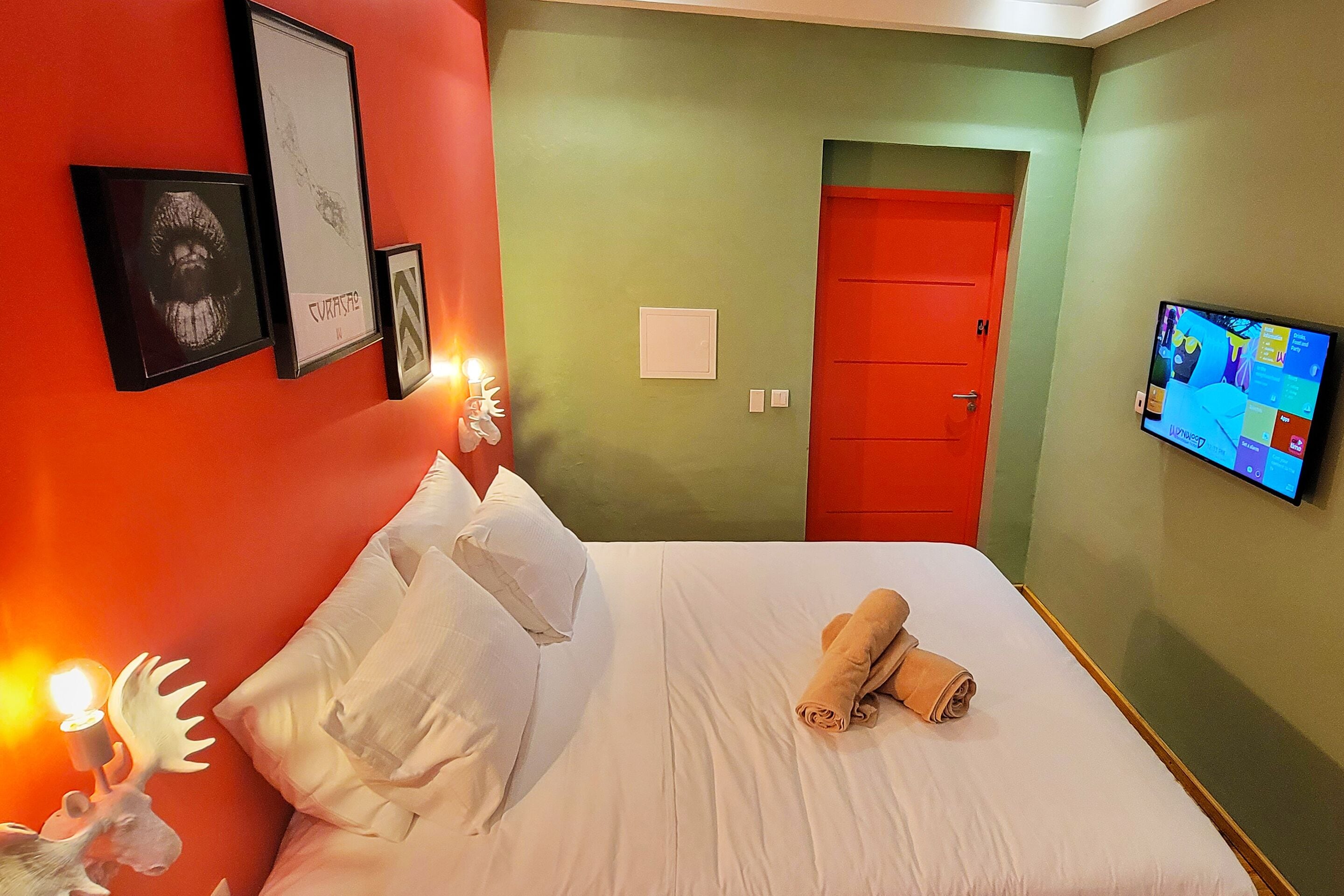 Habitación de hotel con cama, almohadas, una toalla enrollada, televisor en la pared y cuadros enmarcados.