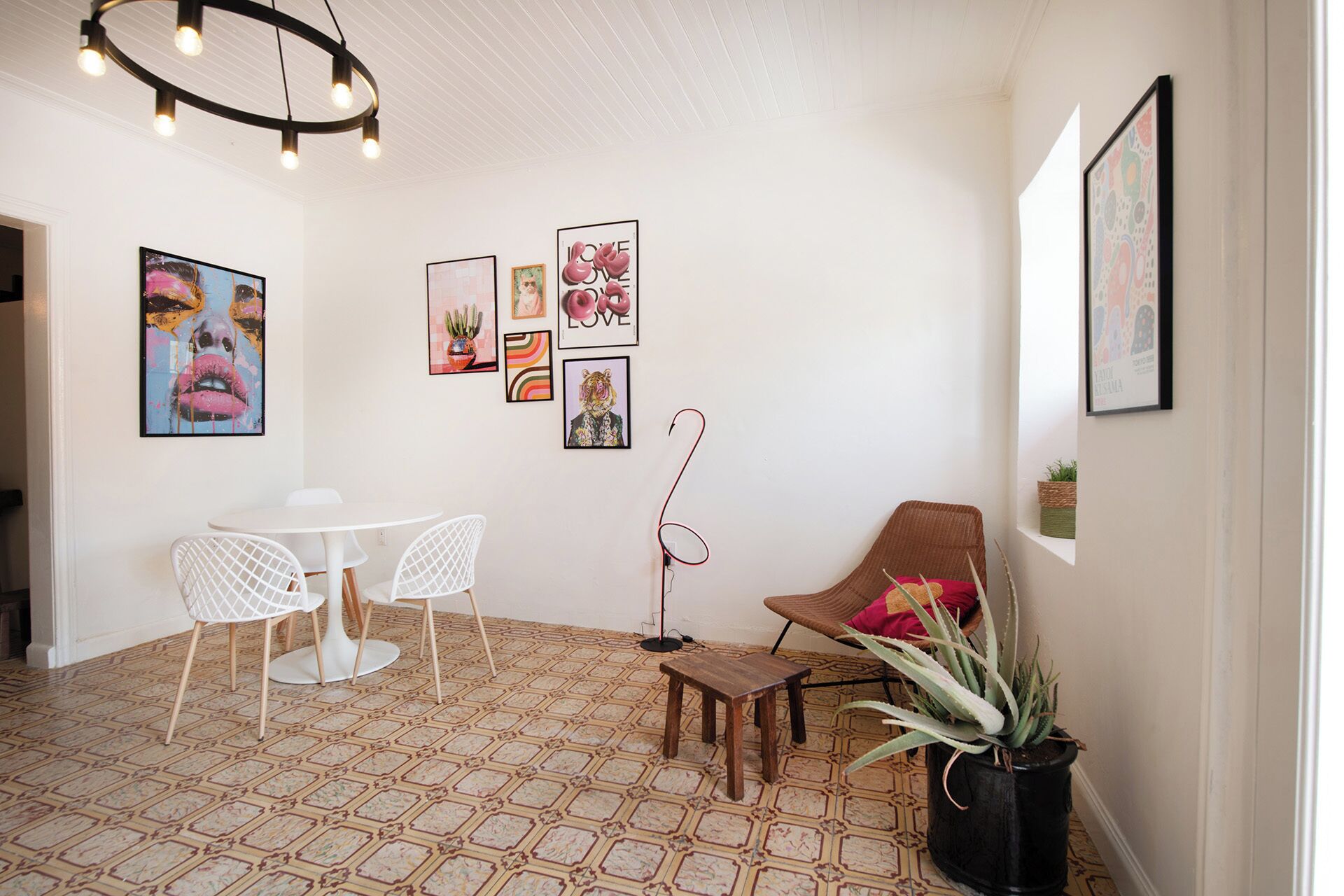 A modern interior with a white ceiling, patterned floor, white chairs, a wooden table, a wicker chair, and various framed artworks on the walls.
