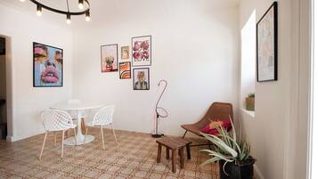 A modern interior with a white ceiling, patterned floor, white chairs, a wooden table, a wicker chair, and various framed artworks on the walls.
