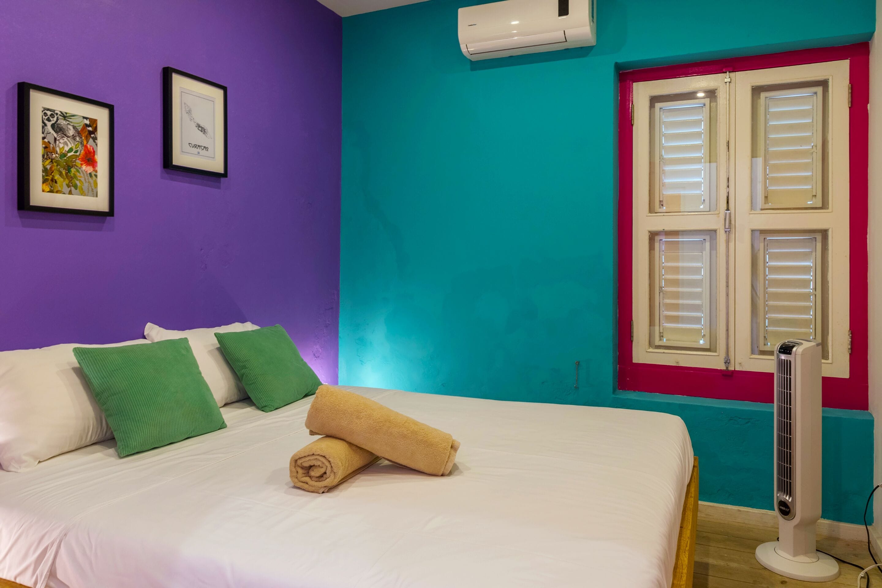 Quarto com cama, travesseiros verdes e brancos, toalha enrolada, ventilador de chão e duas imagens emolduradas na parede.