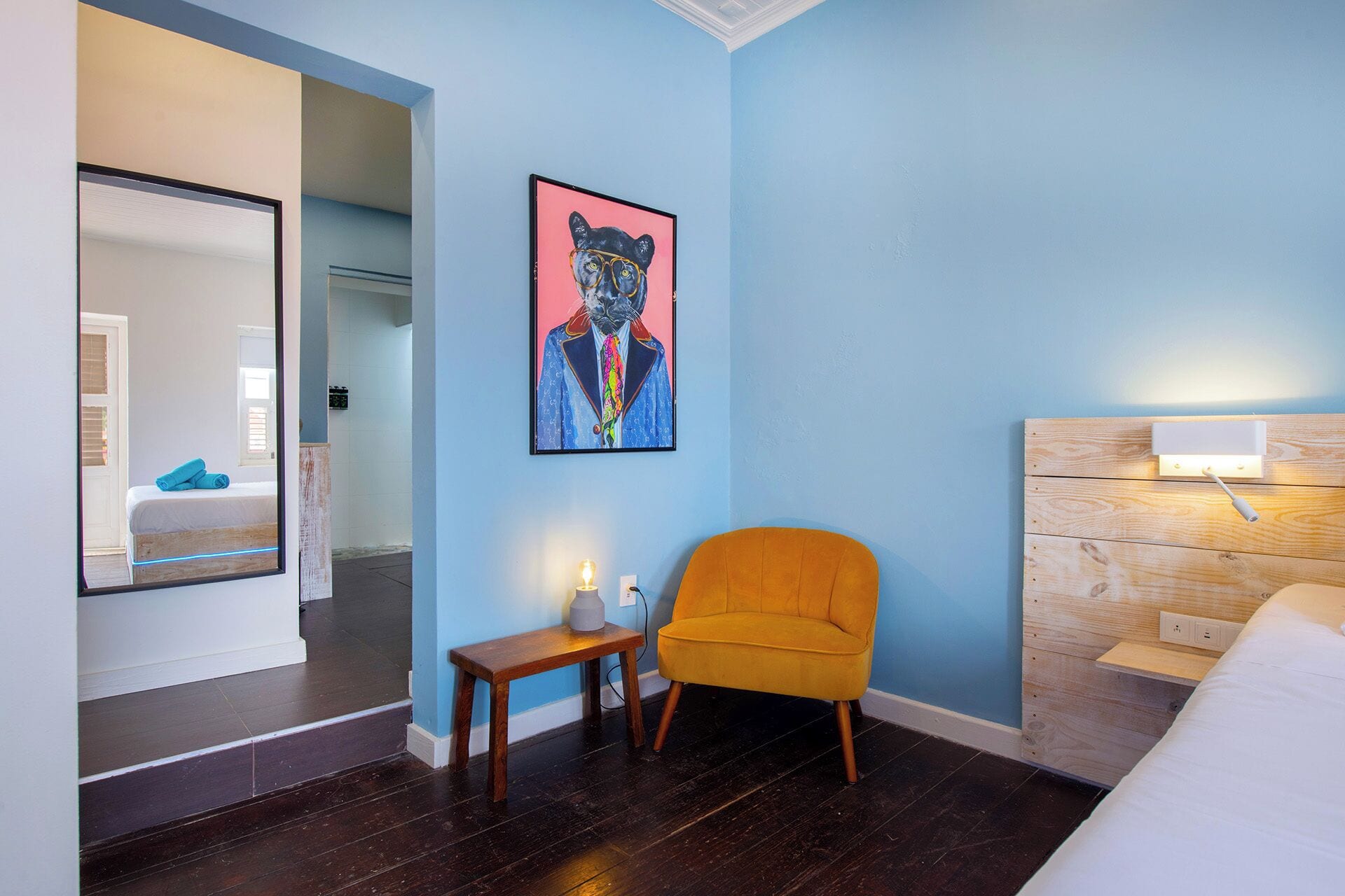 Habitación de hotel con pared azul, cabecera de madera, mesita de noche de madera con una vela y una silla naranja brillante.