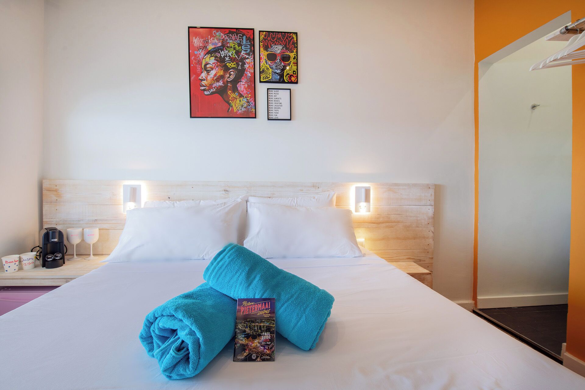 Habitación de hotel con una cama bien hecha, dos obras de arte colgadas en la pared, una cafetera y un libro sobre la cama.