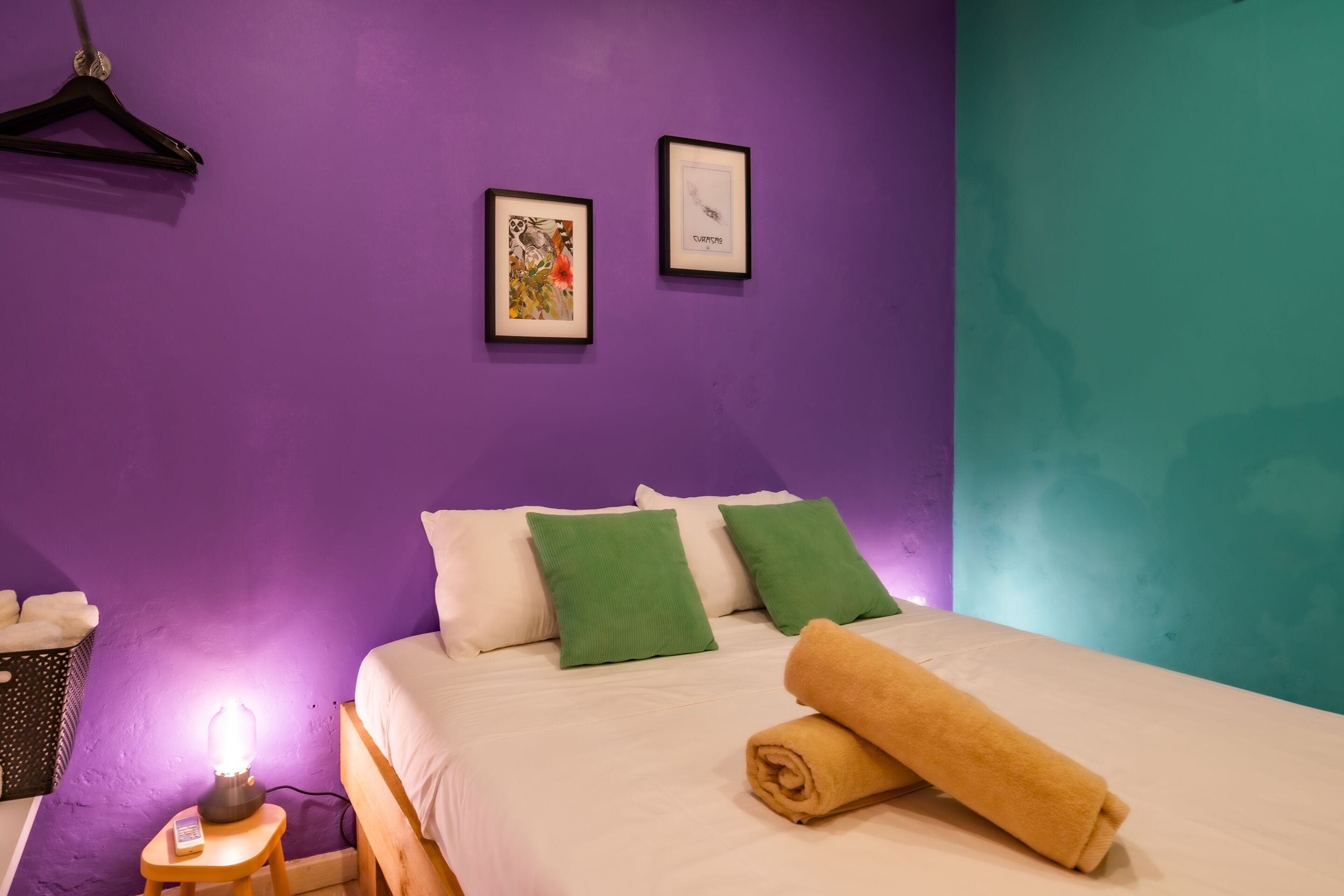 Una habitación con una cama, almohadas verdes, una toalla enrollada, una mesita de noche con una lámpara y dos cuadros en la pared.