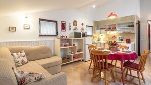 Ein gemütliches Wohnzimmer mit einer Couch und Sesseln, einem Esstisch für vier Personen und einer Küchenzeile mit Mikrowelle, Toaster und Regalen mit Geschirr.