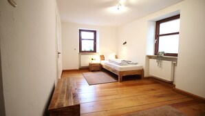 A small, well-lit bedroom with a wooden floor, a single bed, a nightstand, a window, and a radiator.