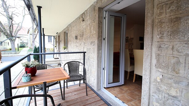A balcony with a wooden table and two wicker chairs, a potted plant, and a sliding glass door leading to an interior space.