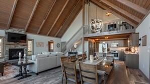 A spacious living area with a fireplace, a dining table with chairs, a staircase leading to an upper level, and a kitchen area.