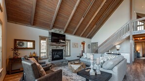 A spacious living room with a stone fireplace, a large television, and a staircase leading to an upper level.