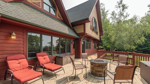 A wooden deck with outdoor furniture, a fire pit, and a view of a house with large windows and a red exterior. - Hot Tub, Fire Pit & Theater Room by Deep Creek (McHenry)