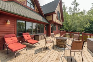 Une terrasse en bois avec des meubles d’extérieur, un foyer et une vue sur une maison dotée de grandes fenêtres et d’une façade rouge.