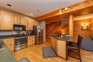A kitchen with wooden cabinets, stainless steel appliances, a dining area with a table and chairs, and a wooden staircase. - Lakefront Bliss: Hot Tub, Views & Fun (McHenry)