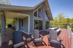 A covered deck with Adirondack chairs and a table, overlooking a house with large windows and a deck.