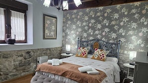 A bedroom with a stone wall, a bed with floral wallpaper, a chandelier, and a bench. - Las Hortensias Country House in Hoznayo (Santa Marina (Entrambasaguas))