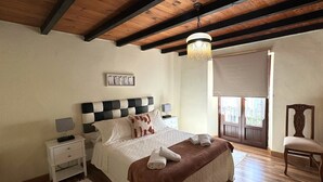 Une chambre avec une tête de lit à carreaux, une table de chevet avec une lampe, un grand lit recouvert d’une couverture marron, un plancher en bois et une chaise en bois.