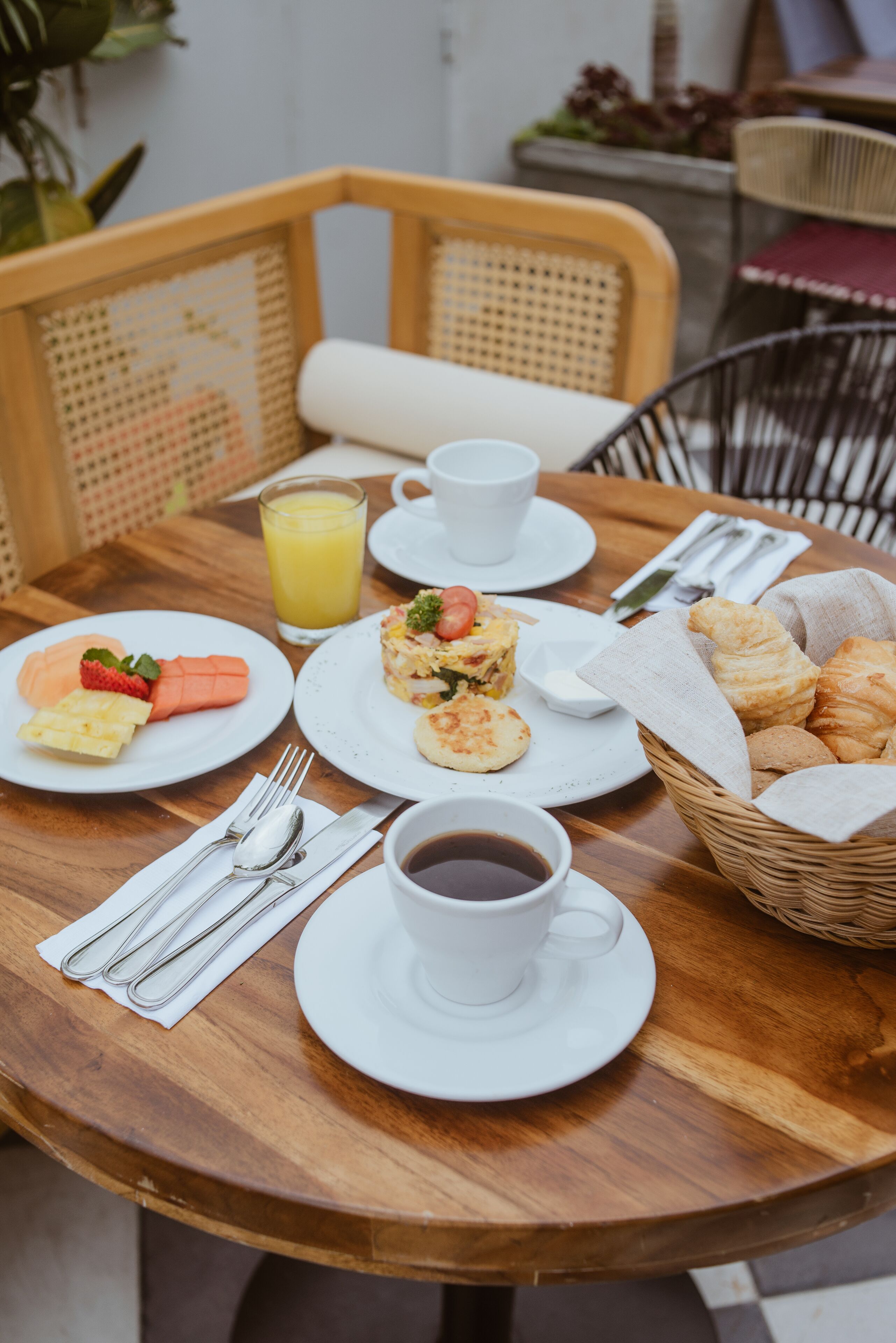 Et træbord med morgenmadsprodukter, heriblandt croissanter, et fad med frugt, et glas appelsinjuice og en kop kaffe.