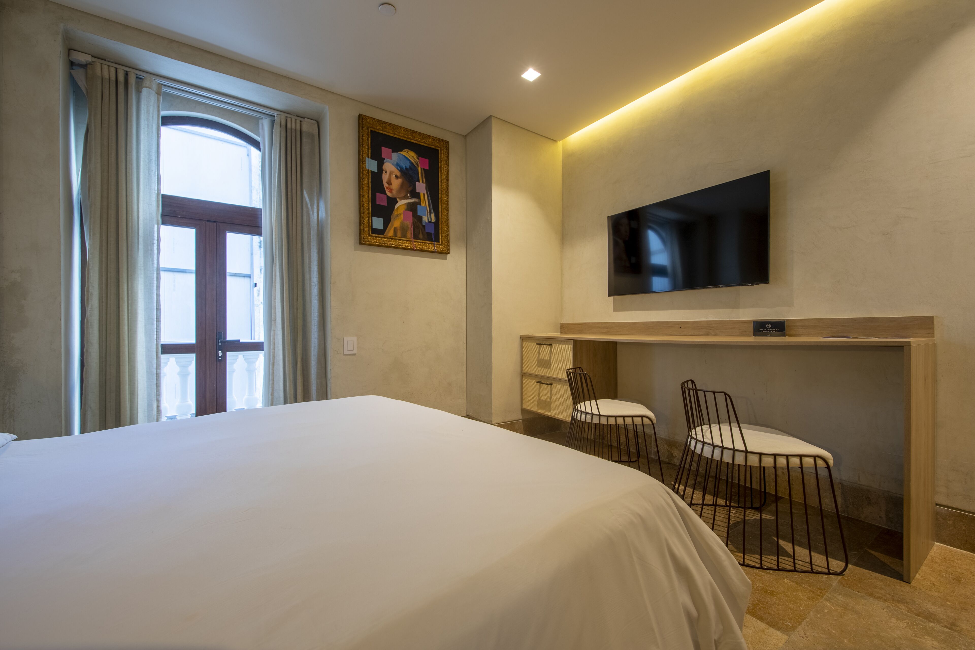 A hotel room with a large bed, a desk with two chairs, a television mounted on the wall, and a painting above the desk.