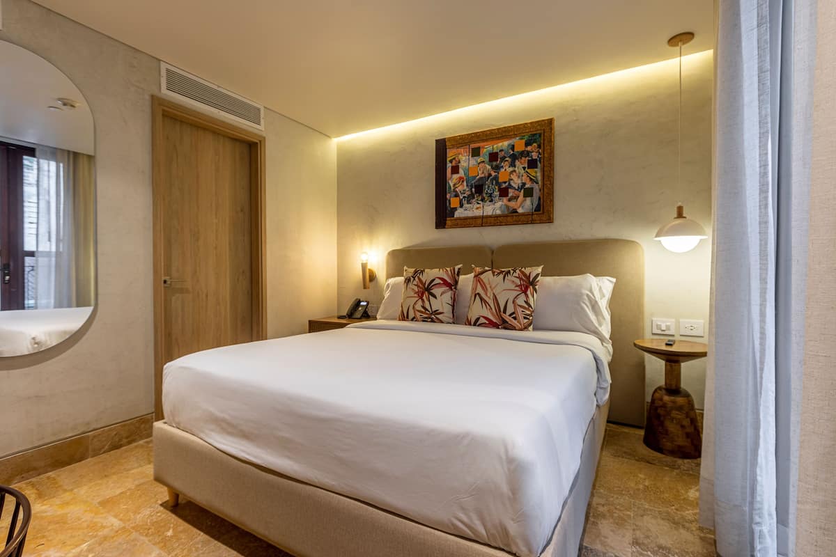 Ein ordentlich eingerichtetes Hotelzimmer mit einem großen Bett, einem Nachttisch mit Lampe, einem Wandbild und einem runden Spiegel.