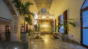 A spacious indoor area with a central seating area, surrounded by tables and chairs. There are large potted plants and a brick wall on one side. - Hotel Voilá Centro Histórico (Cartagena)