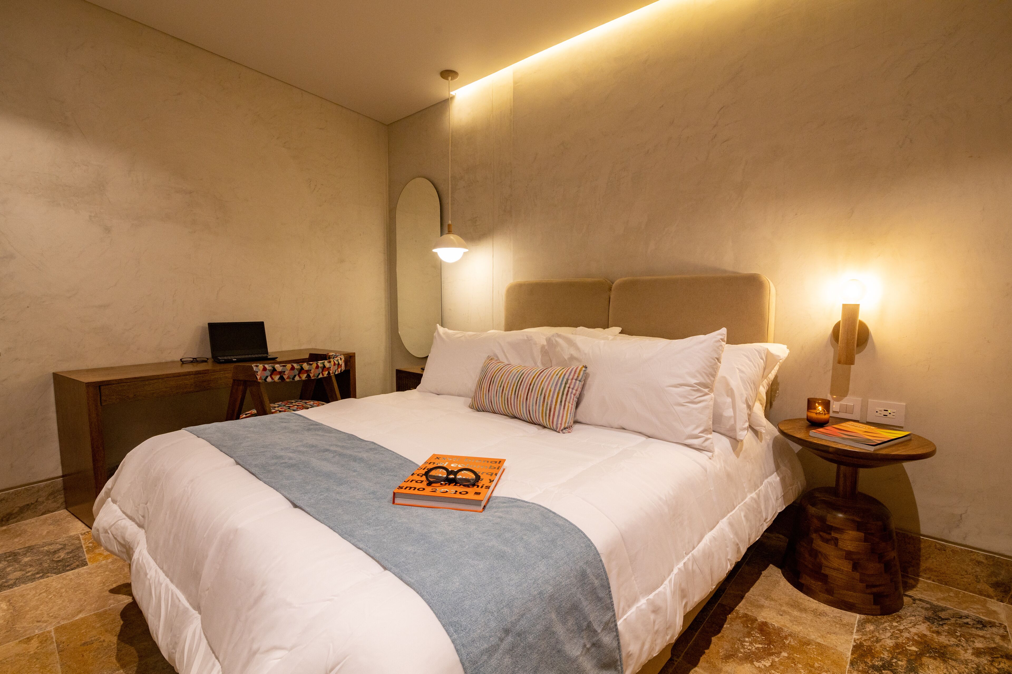 A neatly made bed with a book and glasses, a desk with a lamp, and a small table with a book and candle.
