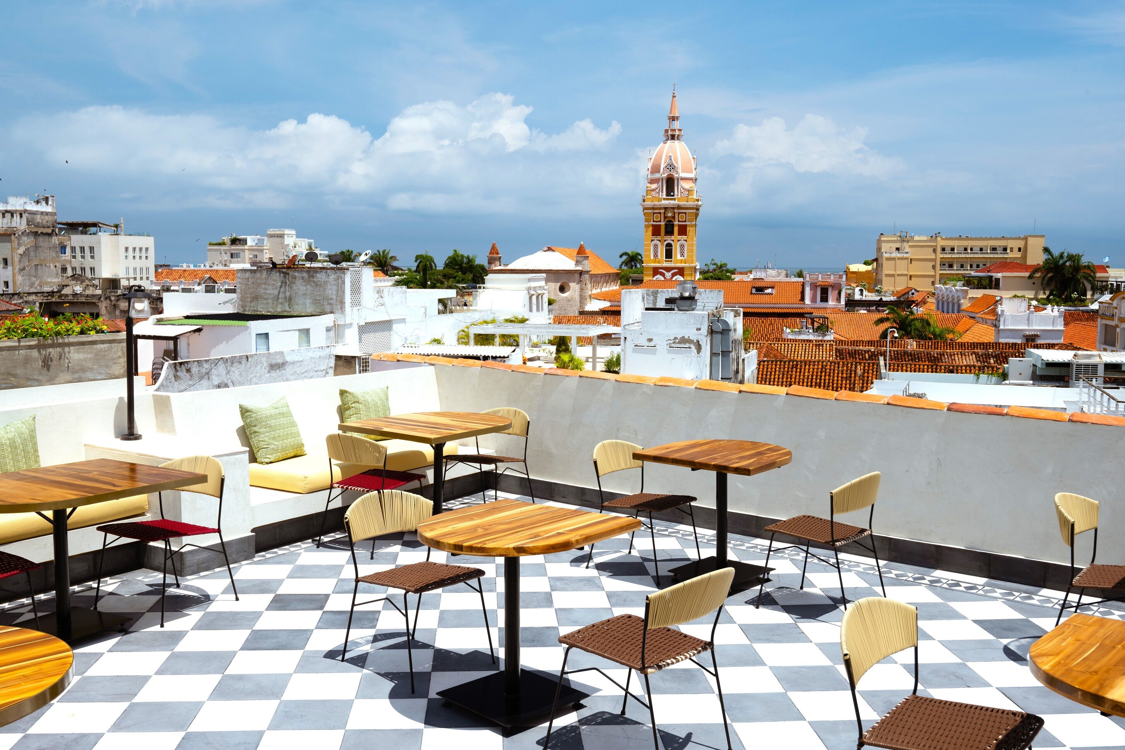 A rooftop terrace with checkered flooring, wooden tables, and chairs, overlooking a cityscape with a prominent tower.