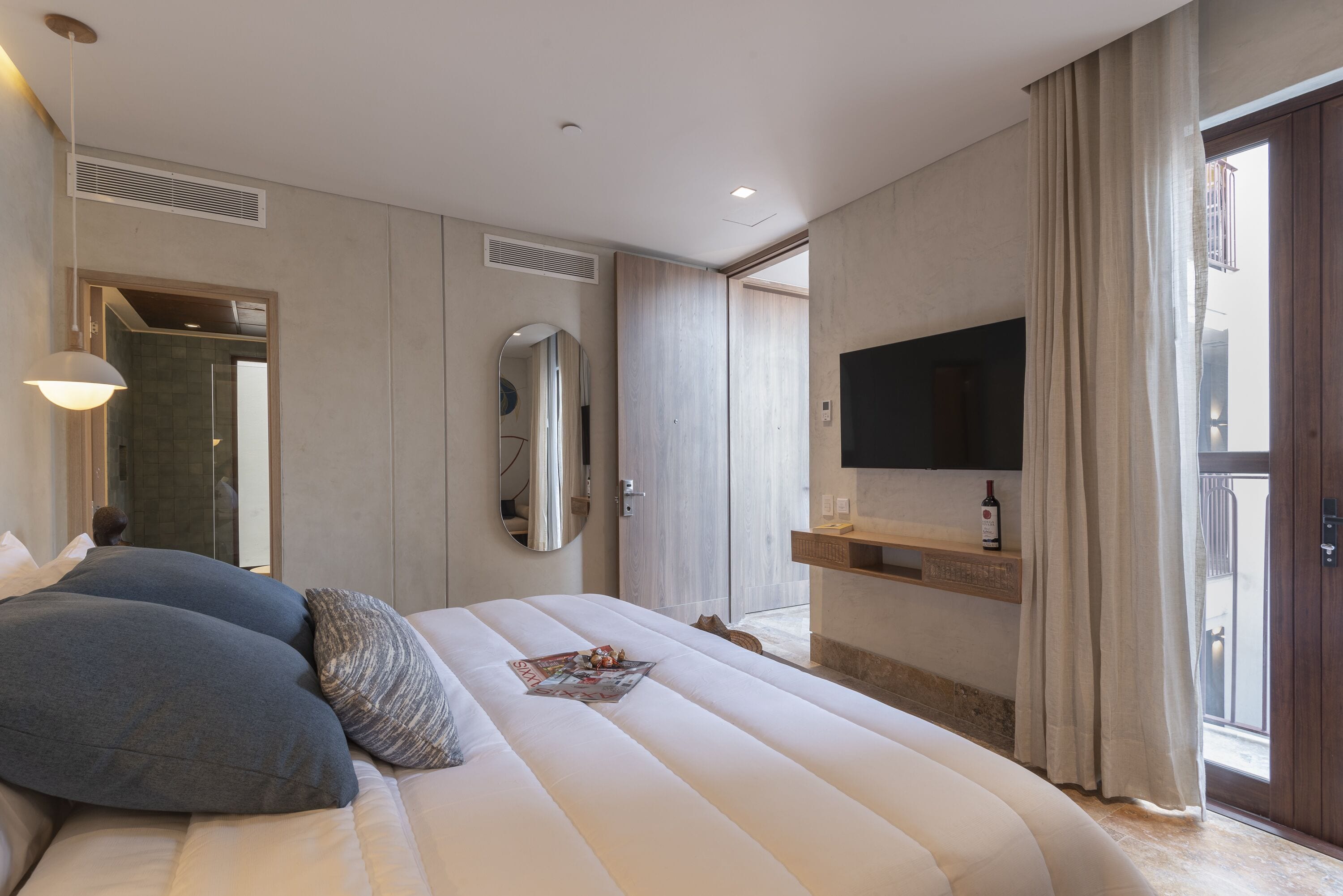 Una habitación de hotel moderna con una cama grande, un televisor montado, un espejo y una ventana con cortinas.