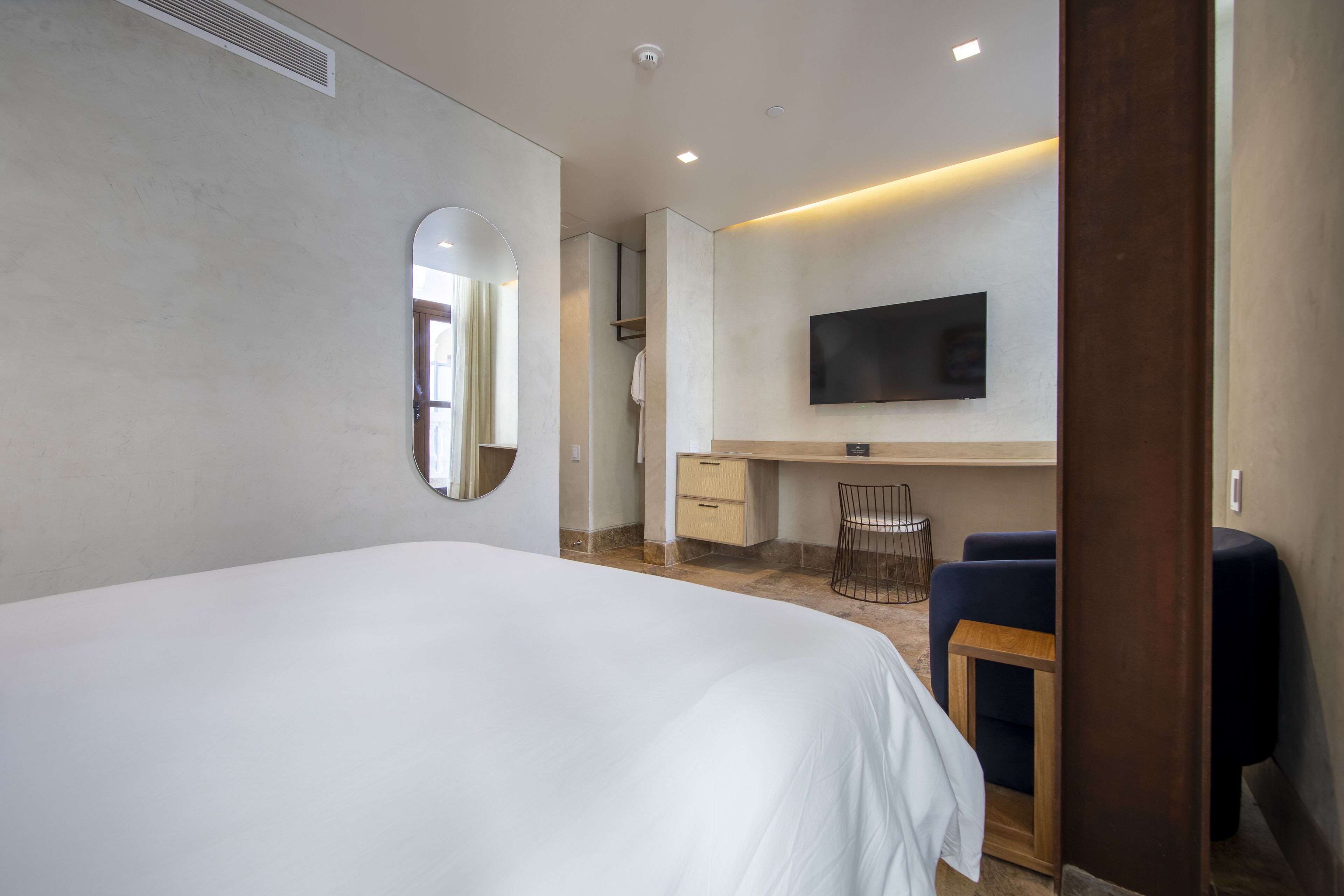 A modern hotel room with a large bed, a desk with a chair, a flat-screen TV mounted on the wall, and a large mirror on the wall.