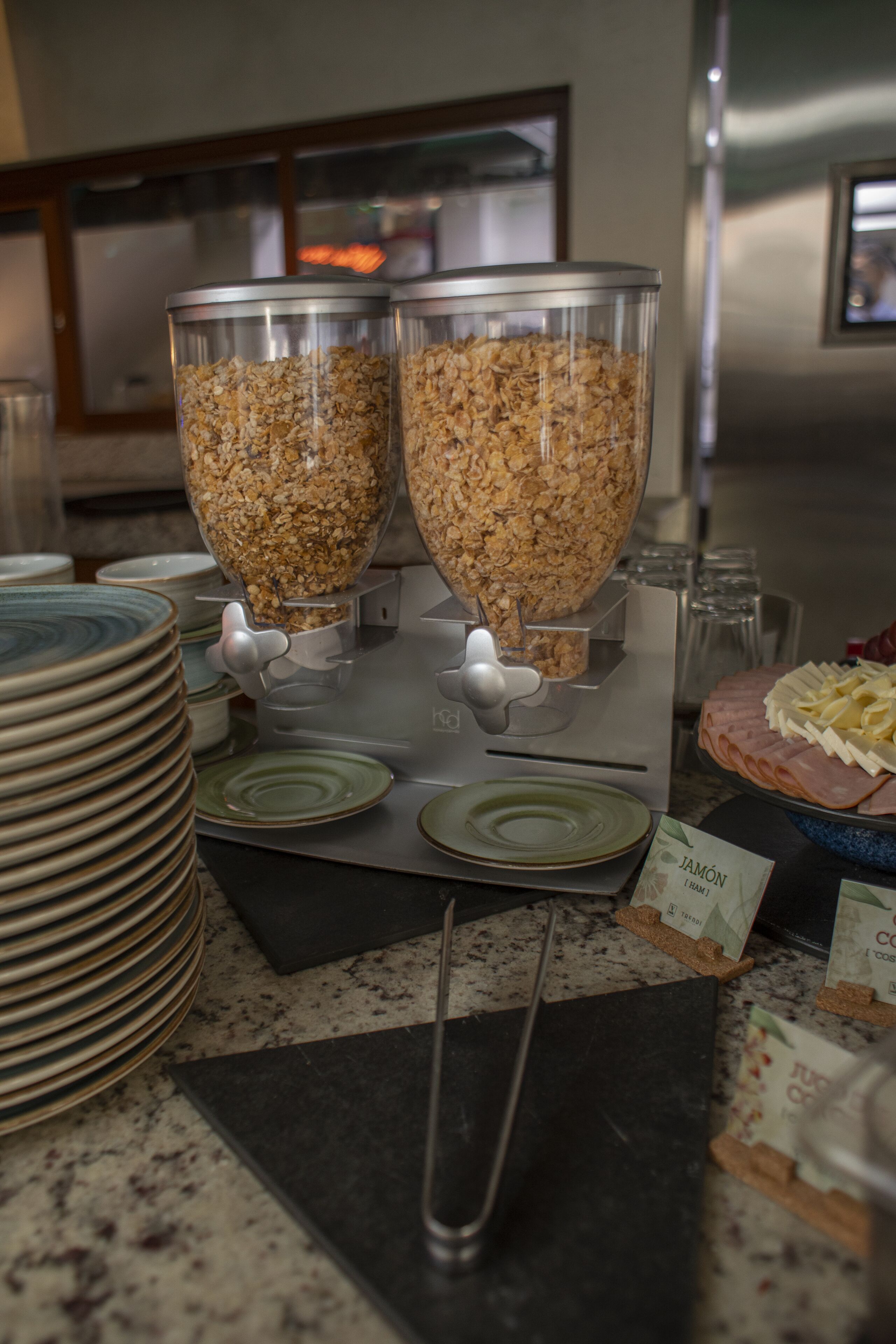 Un espace buffet avec deux grands bols de céréales, une pile d’assiettes et un panneau indiquant une option jambon-fromage.
