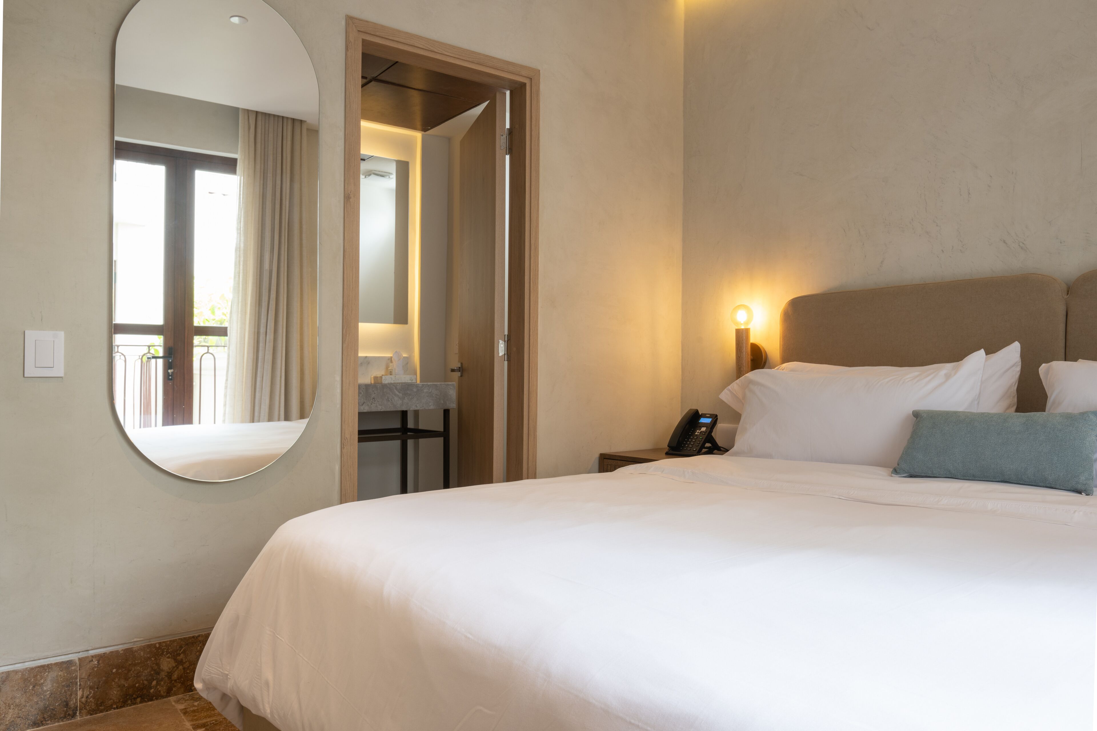 A hotel room with a large bed, a bedside table with a phone, a mirror, and a view of the balcony.