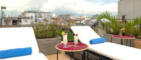 A rooftop terrace with lounge chairs, a small table with drinks, and a city view.