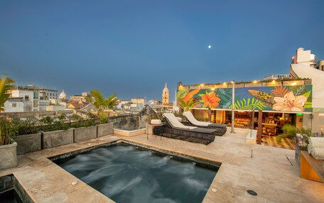 A rooftop pool area with lounge chairs and a hot tub, surrounded by city buildings and a mural.