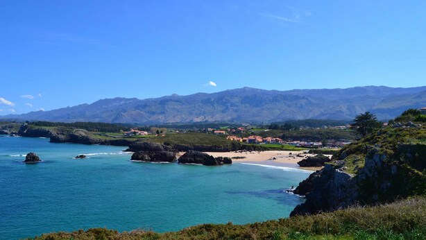 Paesaggio costiero con spiaggia, scogliere rocciose e montagne sullo sfondo.