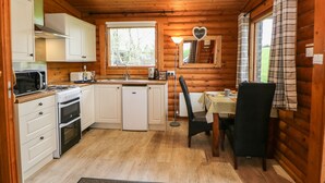 A compact kitchen with white cabinets, a microwave, a stove, and a sink. There's a dining area with a table and chairs, and a window with a view of greenery. - Alwinton Lodge (Morpeth)