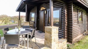 A log cabin with a patio area featuring a round table and wicker chairs, stone pillars, and a view of the countryside. - Alwinton Lodge (Morpeth)
