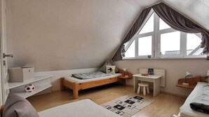 Una habitación pequeña y luminosa, con techo inclinado, una cama, un escritorio y un ventanal con cortinas.