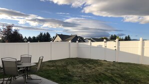 Een achtertuin met een witte hek, een betonnen terras en een eettafel met stoelen.